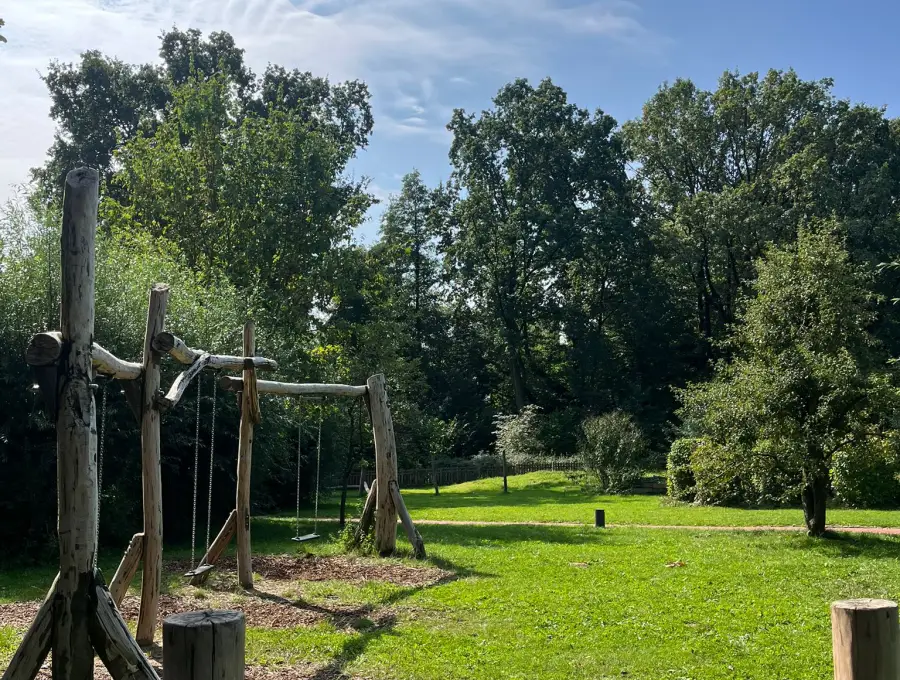 Ein Klettergerüst aus rohen Baumstämmen mit Schaukel auf der Wiese, im Vordergrund eine niedrige Kletterstange