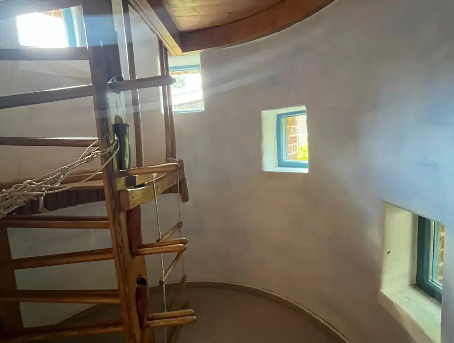 Blick in einen Turmraum mit runder heller Wand, kleinen bunt gerahmten Fenstern und einer Holzleiter, die zu einer Empore hinaufführt