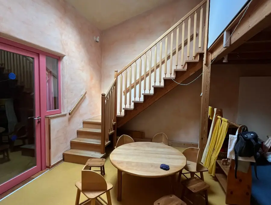 Ein Gruppenraum mit rundem Holztisch und Kinderstühlen, dahinter eine Holztreppe hinauf zur Empore, von oben fällt Licht durch ein Dachfenster