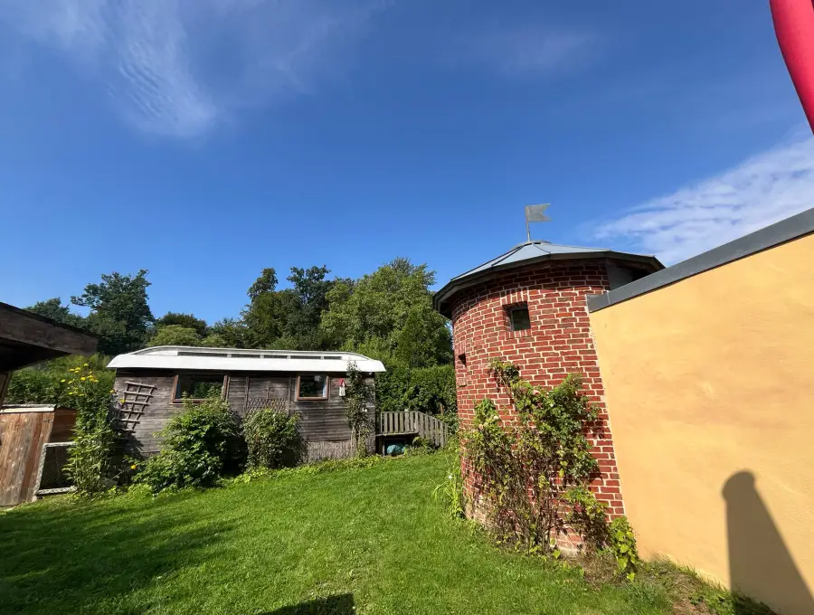 Ein verwitterter Bauwagen aus Holz auf der Wiese, daneben der Backsteinturm mit Wetterfahne
