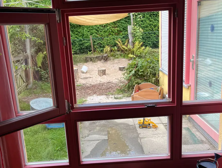 Blick aus einem geöffneten Fenster mit rotem Rahmen auf den Sandspielplatz mit Baumstümpfen und Sonnensegel